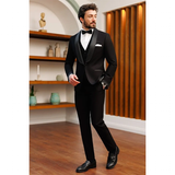 A man in a black tuxedo with a bow tie stands confidently on a wooden floor. He looks to the side with his hand in his pocket, exuding elegance.