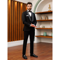Man in a black tuxedo with a bow tie stands on a wooden floor, hands clasped. Wooden panels and decorative shelves in the background create an elegant setting.