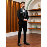 Man in a black tuxedo with a bow tie stands on a wooden floor, hands clasped. Wooden panels and decorative shelves in the background create an elegant setting.