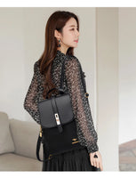 A woman wearing a black floral blouse and light skirt carries a sleek black backpack, showcasing elegant jewelry and hair.