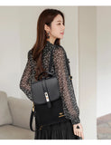 A woman wearing a black floral blouse and light skirt carries a sleek black backpack, showcasing elegant jewelry and hair.