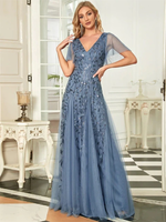 A woman in a blue, floor-length evening gown with floral embroidery poses confidently in a stylish room. The setting has elegant antique furniture.