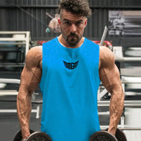 A muscular individual in a bright blue tank top stands in a gym, holding dumbbells, with weightlifting equipment in the background.