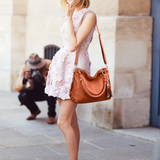 A fashionably dressed woman in a floral pink dress poses with a brown handbag while a photographer captures the moment.