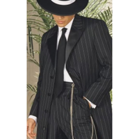 A person wearing a classic black and white pinstripe suit with a tie and a fedora. A chain hangs from their waist. Background has green plants.