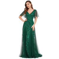 A woman in a floor-length, emerald green gown with a V-neck and sheer leaf-patterned overlay poses confidently, conveying elegance and grace.