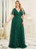 Elegant woman in a flowing green gown with leaf patterns, smiling confidently in a bright room with classical decor and large windows.