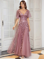 A woman wearing a mauve evening gown with intricate leaf patterns poses gracefully indoors. The dress features a V-neck and sheer sleeves, exuding sophistication.