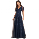 Woman wearing a navy blue evening gown with sheer sleeves and embroidered details. She smiles softly, exuding elegance and confidence against a plain background.