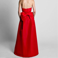 A woman in a strapless red gown with a large bow on the back stands against a plain gray background. The dress exudes elegance and sophistication.
