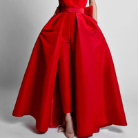 A woman wearing a striking red gown with a voluminous overskirt and tailored pants is standing against a plain gray background. Her posture is poised and elegant.