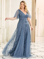 A woman in an elegant, flowing blue gown with intricate leaf patterns stands in a bright room. She looks content, with her arm gracefully extended