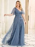 A woman in an elegant, flowing blue gown with intricate leaf patterns stands in a bright room. She looks content, with her arm gracefully extended