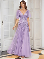 Woman in an elegant lavender gown with leaf patterns, standing in a bright room, smiling softly. The dress has sheer sleeves and a flowing skirt.