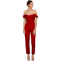 Woman in an elegant red, off-shoulder jumpsuit with ruffle details, poses confidently. She wears nude heels, exuding a sophisticated and chic vibe.