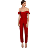Woman in an elegant red, off-shoulder jumpsuit with ruffle details, poses confidently. She wears nude heels, exuding a sophisticated and chic vibe.