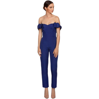 A woman in an off-shoulder, royal blue jumpsuit stands against a white background. She appears poised, gazing to the side, with neutral expression.