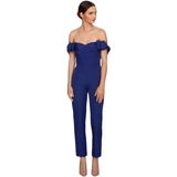 A woman in an off-shoulder, royal blue jumpsuit stands against a white background. She appears poised, gazing to the side, with neutral expression.