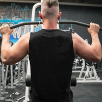 A muscular man in a sleeveless black shirt performs a pull-up, lifting himself on a bar in a gym setting with equipment in the background.