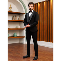Man in a black tuxedo with bow tie stands confidently in a stylish room with wooden floors, shelf décor, and warm lighting, conveying elegance.