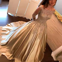 A woman wears an elegant champagne-colored dress with lace detailing and long sleeves, standing in a softly lit room. The gown has a full skirt and a graceful, flowing train.
