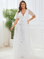 Woman in elegant white gown with floral embroidery stands smiling in bright room. Behind her, a green potted plant and sheer curtains enhance the airy mood.
