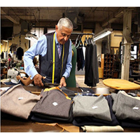 An elderly tailor in a workshop cuts fabric on a table, surrounded by neatly folded clothes. He wears a tape measure and looks focused. The scene conveys craftsmanship.