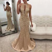 A woman wearing gold sequined evening gown n stands in a room with white walls.She poses elegantly beside a white tufted sofa.