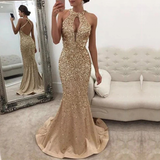 A woman wearing gold sequined evening gown n stands in a room with white walls.She poses elegantly beside a white tufted sofa.