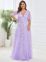 Woman in an elegant, lavender gown with floral patterns smiles in a bright room. The dress features a deep V-neck and sheer short sleeves.