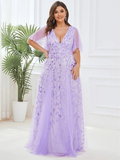 Woman in an elegant, lavender gown with floral patterns smiles in a bright room. The dress features a deep V-neck and sheer short sleeves.