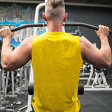 A muscular person in a yellow tank top performs a pull-up in a gym with workout equipment in the background.