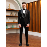 A man in a sleek black tuxedo with a bow tie stands on a wooden floor. The room has shelves with decorative items and a modern, warm ambiance.