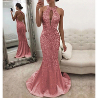 A woman wearing sparkling pink evening gown with a halter neck and open back stands by a mirror. The setting is elegant, with a plush beige sofa.