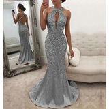 A woman wearing sparkly silver gown stands beside a mirror. The dress features a keyhole neckline and open back. Elegant and glamorous setting.