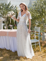 A smiling bride in a white lace gown holds a bouquet beside a table with a checkered cloth. The garden setting is lush and sunny, conveying joy.