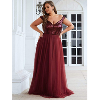 A woman in a burgundy gown stands elegantly on a patio. The dress has a sequined bodice and flowing tulle skirt. She poses with hands on hips, exuding confidence.