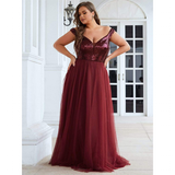 A woman in a burgundy gown stands elegantly on a patio. The dress has a sequined bodice and flowing tulle skirt. She poses with hands on hips, exuding confidence.