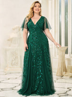 A woman wearing a flowing dark green gown with leaf patterns stands in an elegant room. The dress has sheer sleeves.