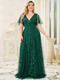 A woman wearing a flowing dark green gown with leaf patterns stands in an elegant room. The dress has sheer sleeves.