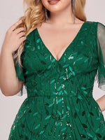 A woman in a green dress with leaf-patterned sequins, featuring a V-neck and sheer sleeves. Her hand touches wavy blonde hair; she wears dangling earrings.