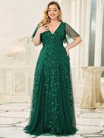 Elegant woman in a luxurious emerald green gown with leaf patterns, standing in a bright, ornate room. She smiles softly, exuding confidence and grace.