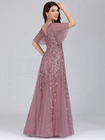 Woman in a mauve evening gown with floral embroidery, sheer short sleeves, and a V-neck back, posing elegantly with a soft smile against a gray backdrop.
