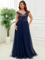 Woman wearing a navy blue, off-the-shoulder, sequined evening gown stands in a bright room with white walls and a potted plant, exuding elegance.