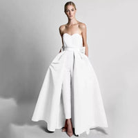 A woman wearing a strapless white gown with a fitted bodice, wide skirt, and slim pants underneath. She exudes elegance against a gray backdrop.