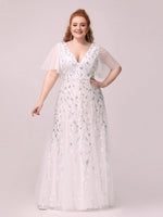 woman in a white, floral-sequined gown with sheer sleeves stands confidently against a light background, embodying elegance and joy.
