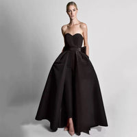 A woman in an elegant black strapless gown with a fitted bodice and full, flowing skirt stands confidently against a plain gray background.