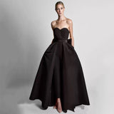 A woman in an elegant black strapless gown with a fitted bodice and full, flowing skirt stands confidently against a plain gray background.