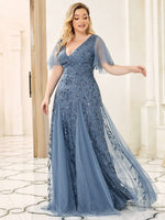 Woman in an elegant blue evening gown with delicate floral embroidery and sheer sleeves, smiles softly in a grand, light-filled room with marble flooring.