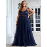 Woman in an elegant, navy blue evening gown with sequined top and flowing tulle skirt, standing confidently by white French doors.